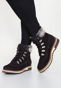 Schwarze Wildleder-Stiefeletten mit grauem Kunstfellbesatz, Schnürdesign, silbernem Zubehör und einer strukturierten Gummisohle mit einem Holzelement.