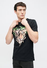 Musta T-paita, jossa on geometrinen kuosi, jossa esiintyy tiikerin kasvot ja vihreää kasvillisuutta. Lyhyet hihat, pyöreä kaula-aukko, sileä puuvillamateriaali.