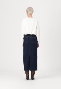 Σκούρα μπλε τζιν φούστα με ξαφρισμένο πίσω μέρος, ψηλή μέση και δύο πίσω τσέπες. Συνδυάζεται με ένα λευκό πουκάμισο μακρύ μανίκι και καφέ μποτάκια με τακούνι.