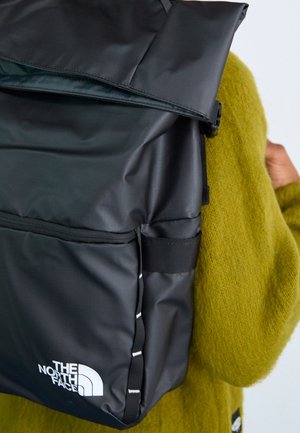Person mit einem schwarzen The North Face Rolltop-Rucksack über einem grünen, strukturierten Pullover, von hinten und seitlich gezeigt.