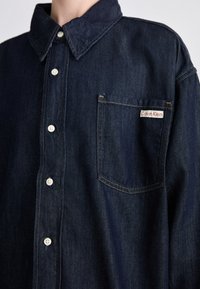 Mørk denim skjorte med knapper og spiss krage, hvite knapper og en brystlomme med en rød og hvit «Calvin Klein»-lapp.