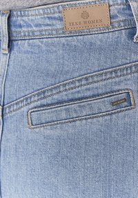 Poche arrière en jean denim bleu clair avec étiquette de marque en cuir beige mentionnant "IKKS Women", cousue au-dessus de la poche.