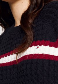 Neulepusero, jossa on tummansininen pohja, ja se on varustettu kaapelitekstuuriin. Punaisia ja valkoisia vaakaraitoja, joissa on pieni logoyksityiskohta raidoissa.