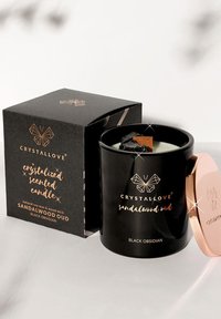 Bocal en verre noir avec un couvercle en rose doré, rempli de cire crème et équipé d'une mèche en bois. La boîte présente un texte noir et doré avec un design géométrique.