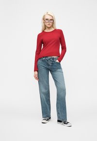 Νεαρή γυναίκα με ξανθά μαλλιά, φορώντας κόκκινο μακρυμάνικο τοπ, μπλε τζιν με κορδόνια, μαύρα αθλητικά παπούτσια και σκούρα γυαλιά ηλίου, στέκεται μπροστά σε λευκό φόντο.