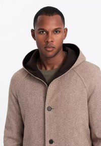 Abrigo texturizado beige con capucha oscura, presenta cierre de botones y mangas raglán, usado sobre un suéter verde. Elementos de diseño limpios y modernos.