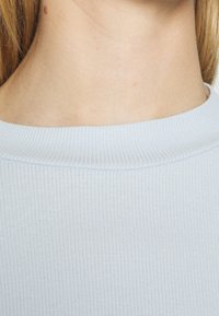 Ljusblått ribbat topp med rund halsringning. Tyget har en slät yta och en subtil lyster som framhäver det ribbade mönstret runt halsen.