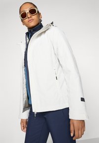 Persoon in een te grote witte hoodie, een marineblauwe ritsjas, bijpassende broek en grote witte zonnebril, tegen een eenvoudige achtergrond.