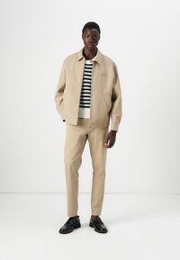 Beige jacka med krage, dragkedjestängning och sidofickor, i kombination med en randig stickad tröja och matchande beige byxor. Svarta skor.