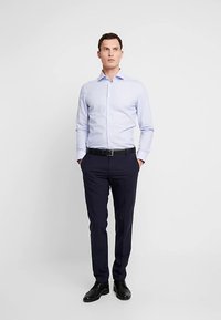 Camicia a righe azzurro chiaro con chiusura frontale a bottoni, abbinata a pantaloni blu navy scuro e scarpe nere. L'ensemble presenta una vestibilità sartoriale.