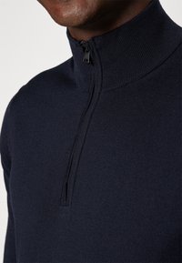 Homme portant un pull en maille bleu marine avec un col montant et une fermeture éclair noire partiellement ouverte au niveau du cou.