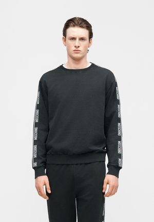 Młody mężczyzna ubrany w czarną bluzę Moschino i dopasowane spodnie z taśmą z logo na rękawach i bokach spodni, stojący na jednolitym tle.