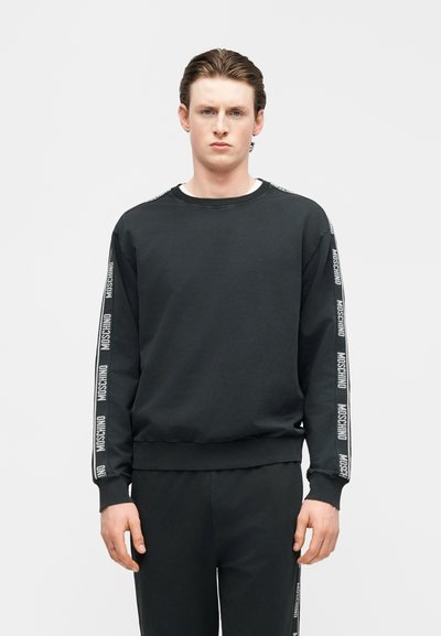 Νεαρός άνδρας που φοράει μαύρο φούτερ Moschino και αντίστοιχο παντελόνι με ταινία λογότυπου στα μανίκια και τις πλευρές του παντελονιού, όρθιος μπροστά σε απλό φόντο.
