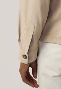 Lichtbeige blouse met lange mouwen, met een knoopsluiting bij de manchetten, gemaakt van een textuurstof en zichtbare stikdetails. Hand ontspannen aan de zijkant.