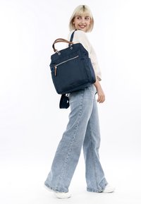 Jeune femme aux cheveux blonds courts portant un jean à jambes larges, des baskets blanches et un pull léger, tenant un grand sac cabas bleu marine.