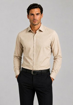 Homme portant une chemise beige boutonnée rentrée dans un pantalon noir avec une ceinture noire, debout les mains dans les poches sur un fond uni.