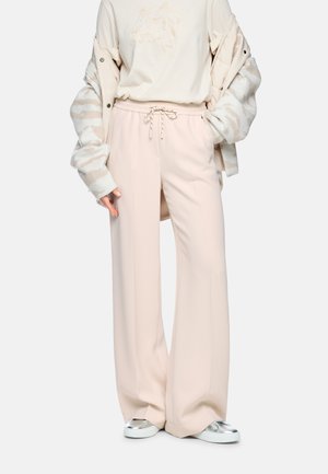 Person wearing wide-leg beige pants with drawstring, a light patterned jacket draped over shoulders, and silver slip-on shoes.