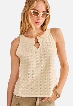 Mujer rubia que lleva un top sin mangas de punto texturizado color crema con escote en forma de ojo de cerradura y gafas de sol beige de estilo ojo de gato, con la mano en el bolsillo.