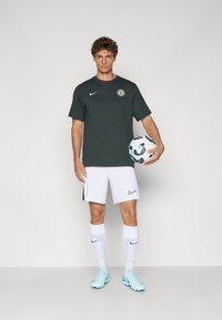 Donkergroene T-shirt met korte mouwen en ronde hals, witte shorts, witte sokken met paarse accenten, en lichtblauwe voetbalschoenen, terwijl hij een voetbals sterkte.