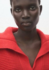 Jersey rojo de punto con un cuello ancho, presenta una textura acanalada. El tono de piel del modelo es profundo, con rasgos faciales definidos y expresión neutral.