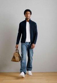 Μαύρο φούτερ με φερμουάρ, λευκό μπλουζάκι, μπλε τζιν και λευκά αθλητικά παπούτσια. Κρατάει μια καφέ δερμάτινη τσάντα. Στέκεται σε ξύλινο πάτωμα ενάντια σε ένα ουδέτερο τοίχο.