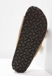 Zapatilla marrón con una suela de goma negra texturizada que presenta un patrón ondulado. La parte superior está hecha de un material suave con un acento esponjoso.