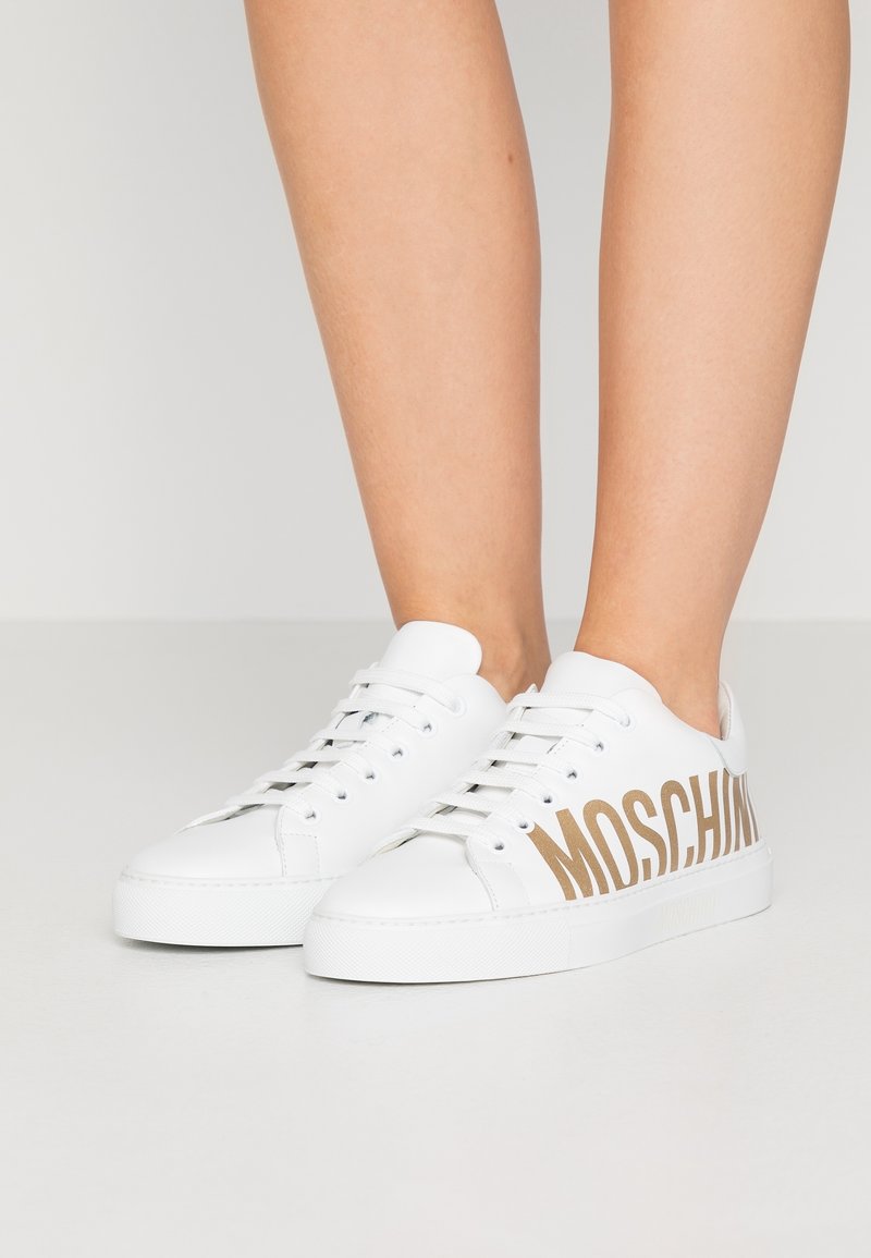 Baskets blanches à lacets avec grand texte doré "MOSCHINO" sur le côté, portées pieds nus sur un fond clair uni.