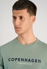 Vihreä puuvillapaita, jossa on pyöreä kaulus, lyhyet hihat ja painettu teksti "COPENHAGEN LINDBERGH" tummansinisellä kirjaimella. Sileä rakenne.