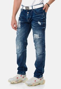 Dunkelblaue Denimjeans mit ausgefransten Stellen und schmalem Schnitt. Sie verfügen über einen weißen Gürtel und sind mit weißen und roten Sneakern kombiniert.