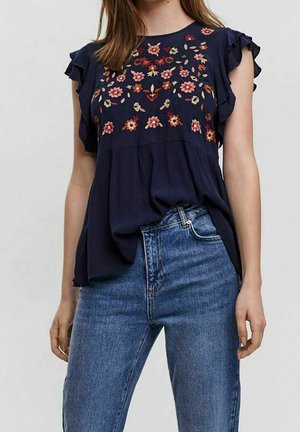 Femme portant un chemisier bleu marine à manches volantées et broderies florales multicolores, associé à un jean bleu taille haute.