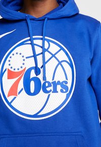 Sweat à capuche bleu présentant un grand logo circulaire blanc avec des accents rouges et bleus, affichant "76ers" et un graphisme de ballon de basket.