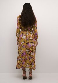 Ilgas, gėlių raštuotas suknelė geltonos spalvos su purpurinėmis ir baltomis gėlėmis. Suknelė turi prigludusį liemenį ir ilgas rankoves, derinama su šlepetėmis ant aukšto kulno.