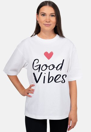 Young woman with long brown hair wearing a white oversized T-shirt with a red heart and "Good Vibes" text, standing with one hand on hip.
