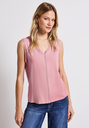 Blouse sans manches rose avec un décolleté en V et un bord en dentelle décoratif sur le devant. Le tissu semble léger et doux. Associée à un jean bleu.