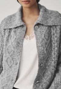 Cardigan grigio lavorato a maglia con ampio colletto, caratterizzato da un motivo testurizzato, grandi bottoni, indossato sopra una maglietta bianca con bordatura in pizzo.