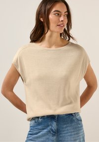 Beige stickad topp med kort ärm, rund halsringning, mjuk textur och avslappnad passform. Kombinerad med blå denim, vilket skapar en avslappnad stil.