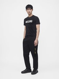 Άνδρας στέκεται φορώντας μαύρο t-shirt Moschino, μαύρο παντελόνι με κίτρινο φερμουάρ και μαύρα αθλητικά παπούτσια σε λευκό φόντο.