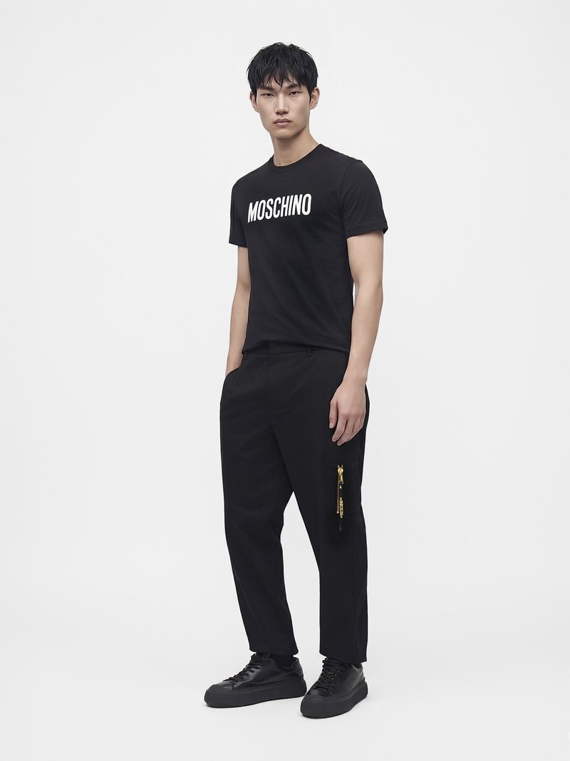 Мъж, облечен с черна тениска Moschino, черен панталон с жълт цип и черни маратонки на бял фон.