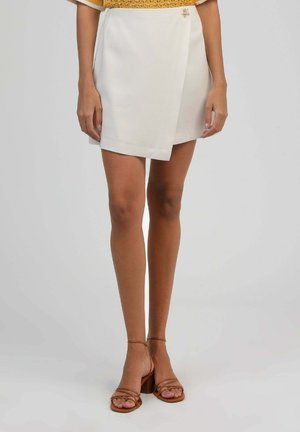 Femme portant une jupe portefeuille asymétrique blanche avec une petite broche fleurie, associée à des sandales à talons blocs marron à brides.