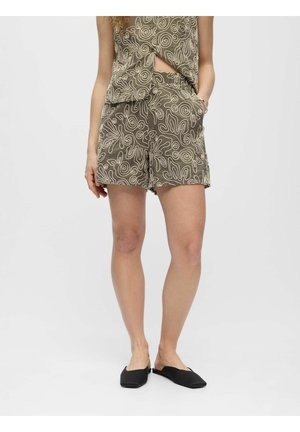 Femme portant un short marron et un haut assorti avec des motifs abstraits blancs en lignes, debout avec une main dans la poche et des chaussures noires sans lacets.