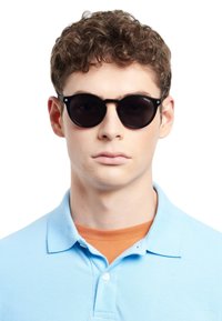 Occhiali da sole neri e rotondi con lenti scure, dotati di una montatura in plastica minimalista. Indossati da una persona in una polo azzurra chiara.