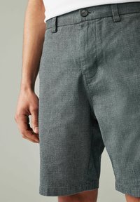 Shorts tissés gris avec une finition texturée, dotés de poches latérales et d'une fermeture à bouton à la taille. Gros plan sur les shorts et une main.