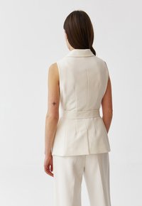 Hihaton kerma värinen liivi, jossa on istuva vyötärö, peplum-muotoilu ja kärkipäinen kaulus. Sujuva pinta, jossa on pystysuorat saumat ja minimalistiset yksityiskohdat.