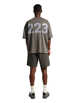 Mężczyzna zwrócony tyłem, ubrany w oversize szary T-shirt z dużym „223” na plecach, ciemne spodenki, białe skarpetki i szare sneakersy na białym tle.