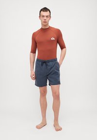 Brun kortermet rash guard med en hvit logo på brystet, kombinert med marineblå svømmebukser med elastisk midje og snøring.