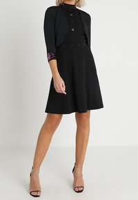 Černé šaty s přiléhavým živůtkem a rozšířenou sukní, zdobené stříbrnými ozdobami, doplněné krátkým černým kardiganem s květinovým motivem na rukávech.