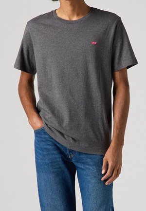 Mand iført en ensfarvet mørkegrå t-shirt med korte ærmer og lille rødt Levi's-logo på brystet, kombineret med blå jeans, hånd i lommen.