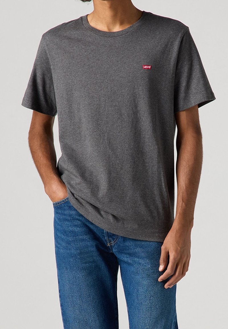 Homme portant un t-shirt à manches courtes gris foncé uni avec un petit logo rouge Levi's sur la poitrine, associé à un jean bleu, main dans la poche.