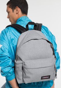 Grauer Stoffrucksack mit einem Hauptfach, Fronttasche, schwarzen Gurten und Eastpak-Logo. Glatte Textur und schlichtes Design.