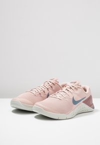 Zapatillas deportivas de color rosa claro con parte superior de malla transpirable, logo de Nike en azul, acentos texturizados en el talón y suela blanca. Diseñadas para ofrecer comodidad y soporte.